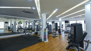 A gym with various exercise machines, a wall-mounted dumbbell rack, and a large mirror. - Spoorti Resort and Club (Bijapur)