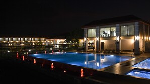 A swimming pool with a water fountain feature, a covered seating area, and a building with large windows and a balcony. - Spoorti Resort and Club (Bijapur)