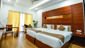 A hotel room with two beds, a desk, a chair, a ceiling fan, and a window with curtains. - Spoorti Resort and Club (Bijapur)