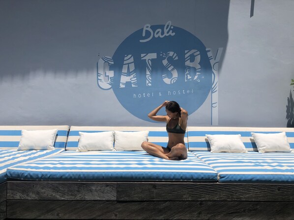 A person sitting on a blue and white striped sofa with white pillows, in front of a wall with a large circular logo and text reading "Bali GATSBY hotel & hostel". - Gatsby Hotel and Hostel (Pecatu)