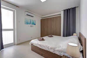 A modern bedroom with a wooden bed frame, a white mattress, and a bedside lamp. There is a window with curtains, an air conditioning unit, and a painting on the wall. - Gorgeous 1BR Apartment close to Amenities (Msida)