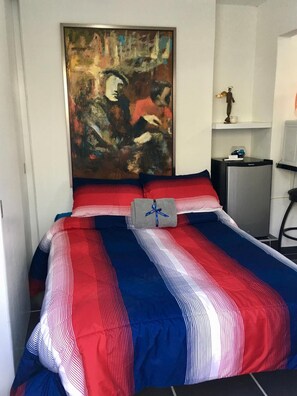 Uma cama com colcha listrada em vermelho e azul, um pequeno travesseiro cinza e uma pintura sobre a cama.