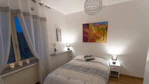 Een slaapkamer met een bed, nachtlampjes, een raam met gordijnen, een radiator en een wand met een kleurrijk schilderij.