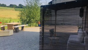 Een terras met een houten tafel en banken, een vlechtwerk privacy scherm, en een geplaveid pad.