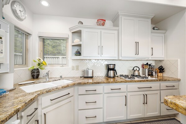 A kitchen with white cabinets, granite countertops, a clock, and a window with a view of trees.