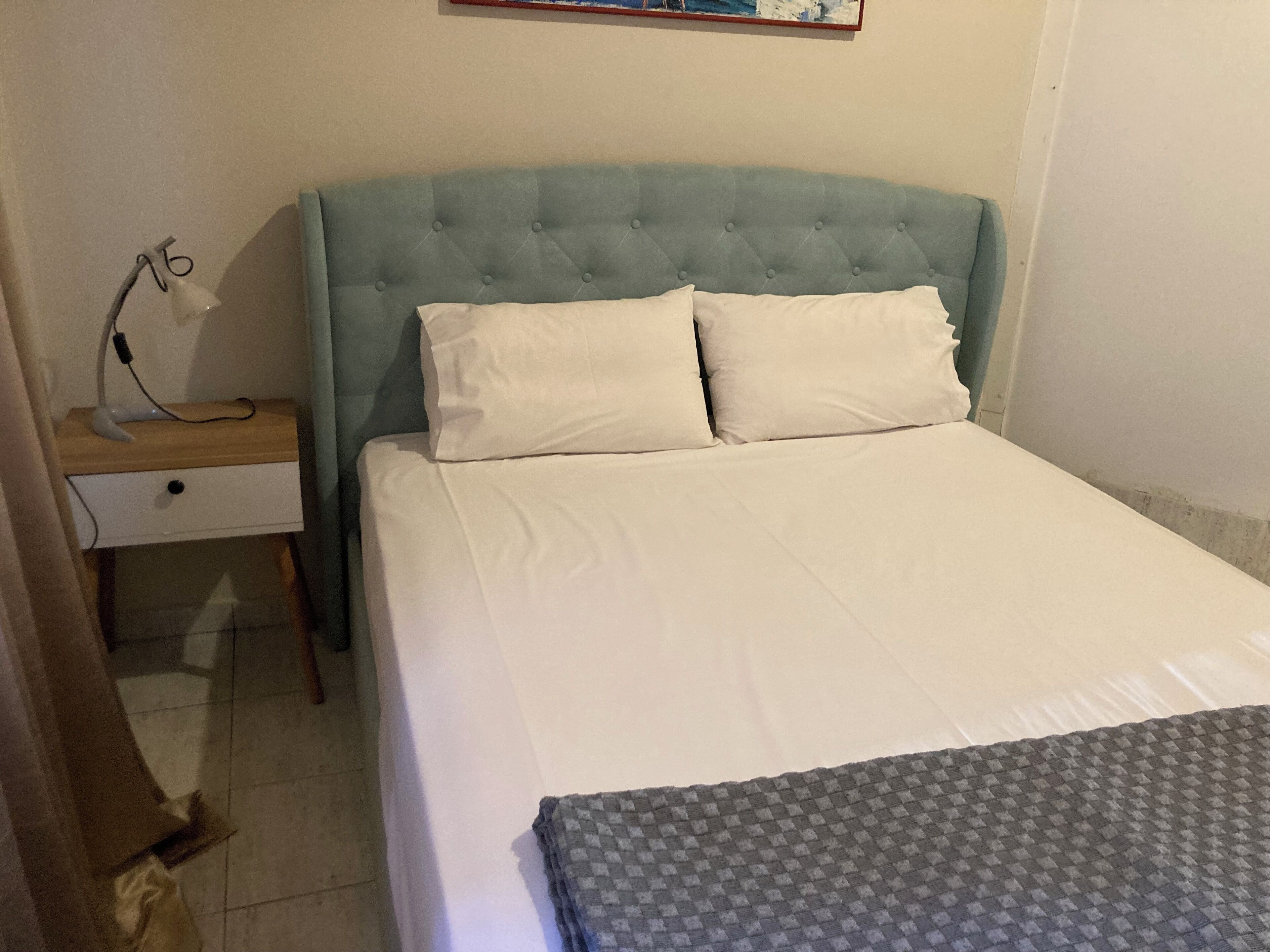 Una cama bien hecha con cabecera capitonada, una mesita de noche con lámpara y un pequeño cajón.
