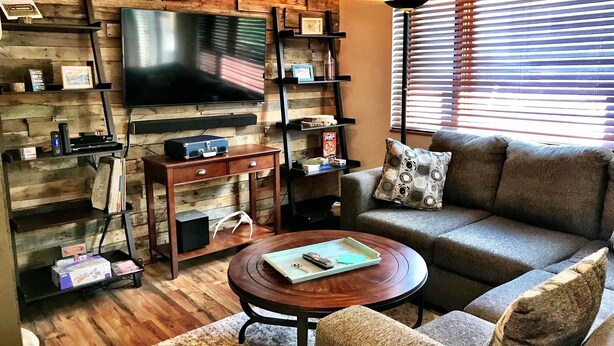 A living room with a wooden wall, a flat-screen TV, a sofa, a coffee table, and a lamp. - Carbondale Townhome with A/C, WiFi, and Backyard (Carbondale)