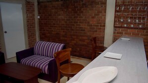 A room with a brick wall, a sofa, a wooden chair, a dining table with a white plate, and a shelf with cups. - Laana Villa (Negombo)