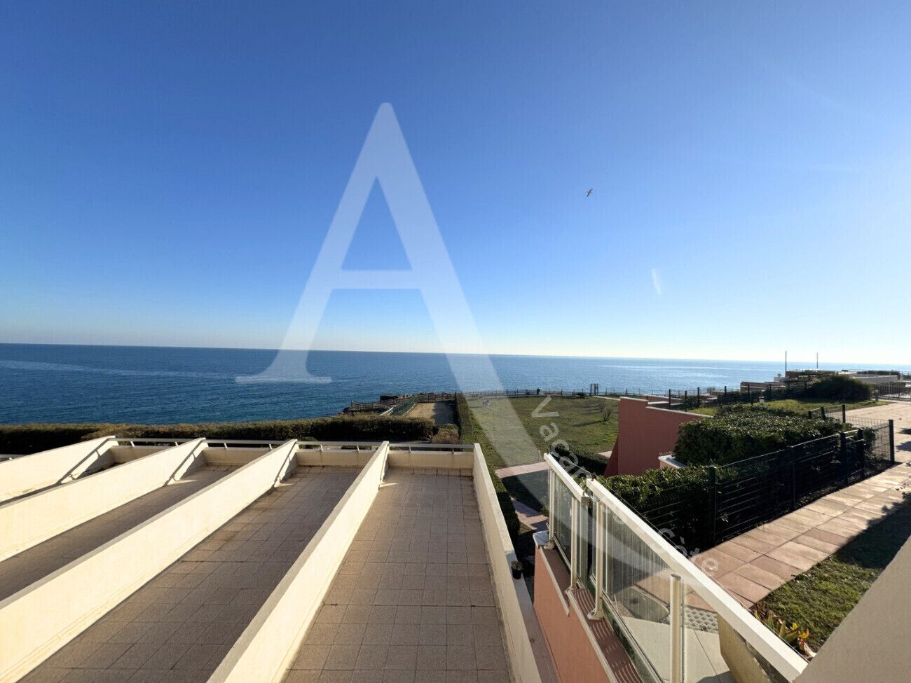 A balcony with a view of the sea, a paved area, and a grassy section with a fence.