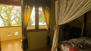 A room with a bed, curtains, a fan, and a window with a view of trees. - Panorama Backpackers (Pulau Bunaken)