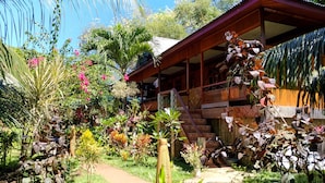 A tropical garden with a wooden house featuring a balcony and a staircase. - Panorama Backpackers (Pulau Bunaken)