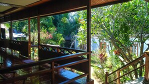 A covered outdoor area with a wooden railing, a tiled floor, and lush greenery visible through the railing. - Panorama Backpackers (Pulau Bunaken)