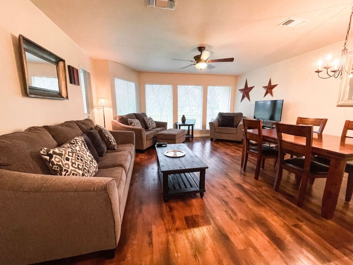 A living room with a sofa set, a coffee table, a dining table with chairs, a ceiling fan, and large windows.