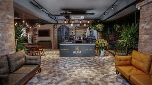 Un vestíbul d'hotel amb una taulell de recepció que diu "ALFA HOSTEL HOTEL", paviment amb dibuixos hexagonals i diverses zones d'estar incloent sofàs i un butac armchair groc.