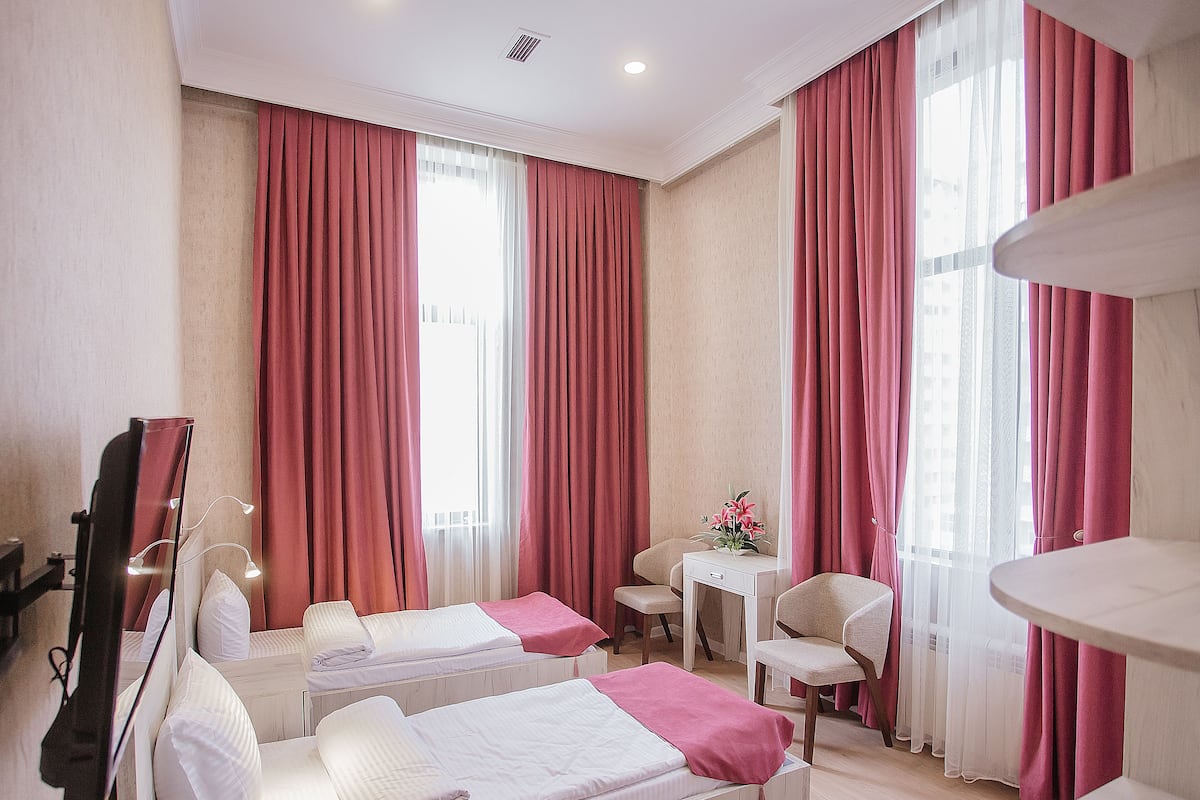 Une chambre d’hôtel avec deux lits, un bureau, une chaise et de grandes fenêtres avec des rideaux rouges.