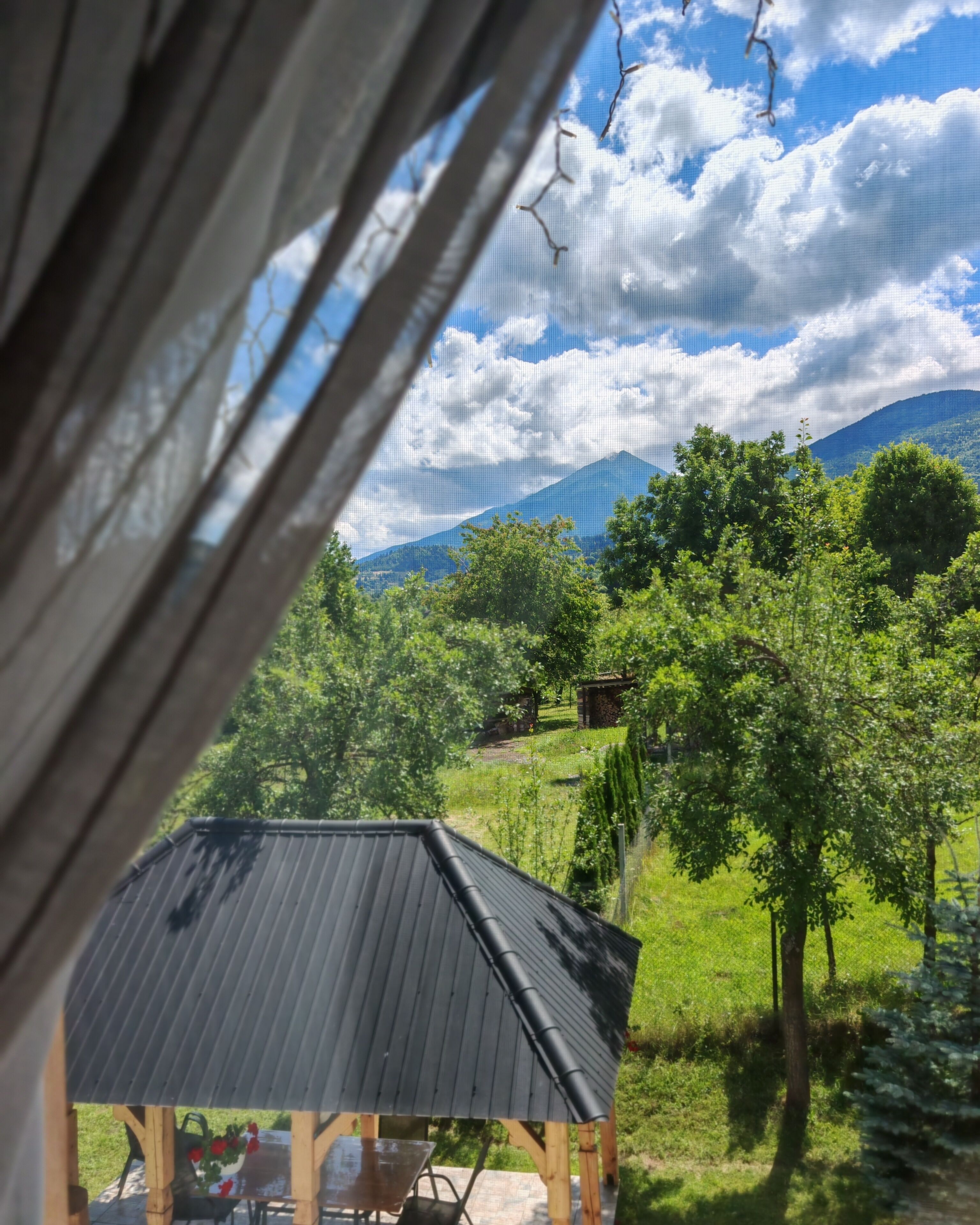 A rustic wooden structure with a sloped roof, surrounded by lush greenery and a mountain in the background.