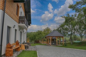 A building with a wooden exterior, a stone bench, a gazebo, and a grassy area. - Pensiunea Maramures Landscape (Moisei)