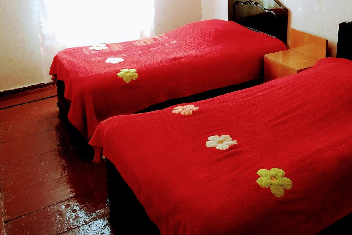 Una habitación con dos camas, cada una cubierta con ropa de cama roja y una sola flor decorativa. Hay una mesita de noche de madera entre las camas y una pequeña mesa de madera junto a ella. Las paredes son blancas y hay una ventana con cortinas de encaje.