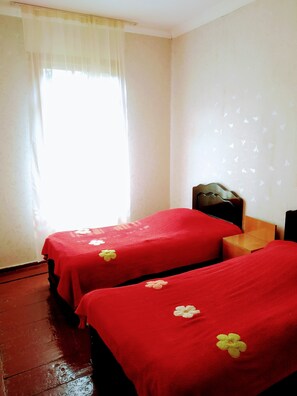 Una habitación con dos camas, cada una cubierta con ropa de cama roja y una sola flor decorativa. Hay una mesita de noche de madera entre las camas y una pequeña mesa de madera junto a ella. Las paredes son blancas y hay una ventana con cortinas de encaje.