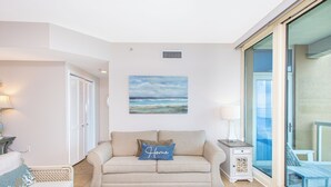 Ein Wohnzimmer mit einem beigefarbenen Sofa, einem blauen Kissen mit dem Aufdruck "Home" und einem Bild von einem Strand. Ein Balkon mit einer Glastür ist ebenfalls zu sehen.