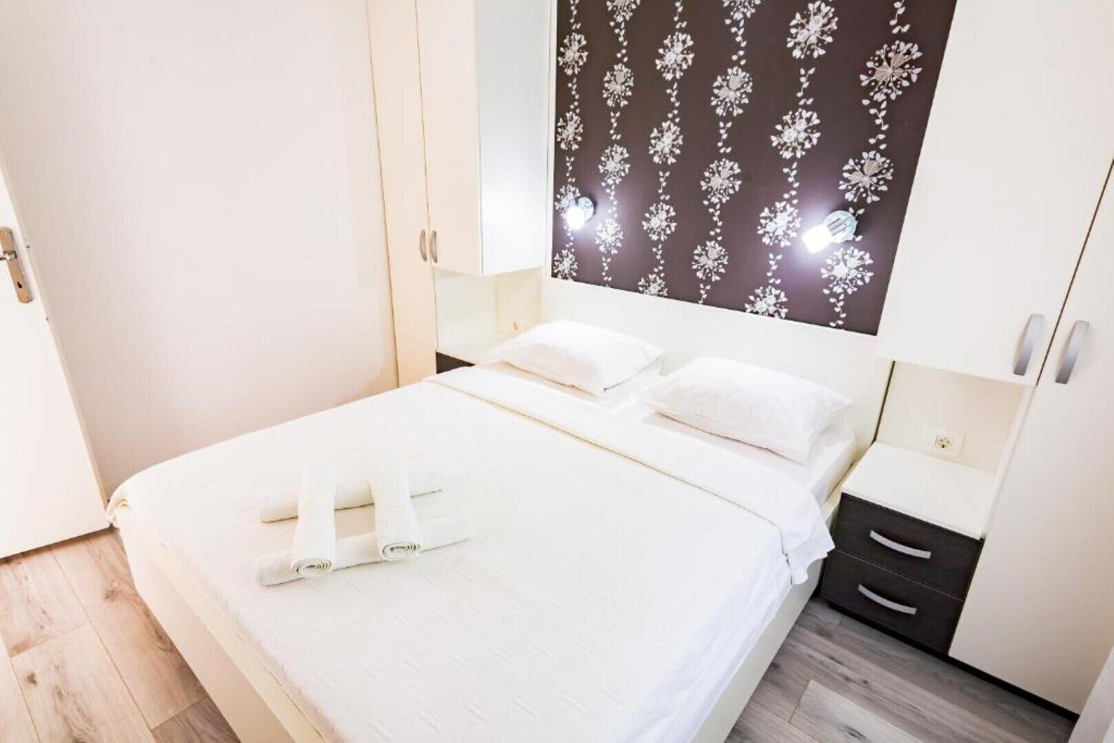 A neatly made bed with white linens, a dark patterned headboard, and a bedside table with a lamp.