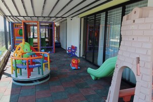 Un área de juegos al aire libre techada con diversas estructuras, como un gimnasio de escalada, un tobogán y una pequeña piscina de pelotas.
