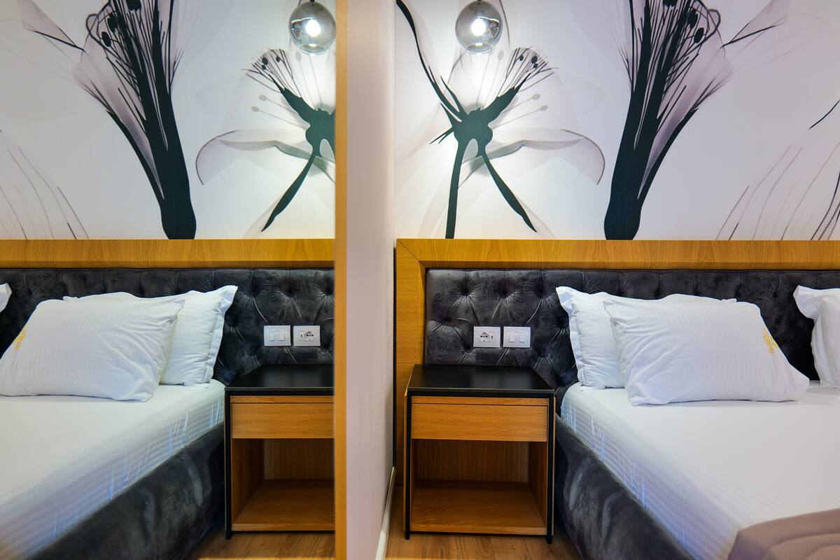 Een hotelkamer met twee bedden, elk voorzien van een houten hoofdbord en wit beddengoed. Er staan twee nachtkastjes met lampen, en de muren zijn versierd met bloemistische kunstwerken.