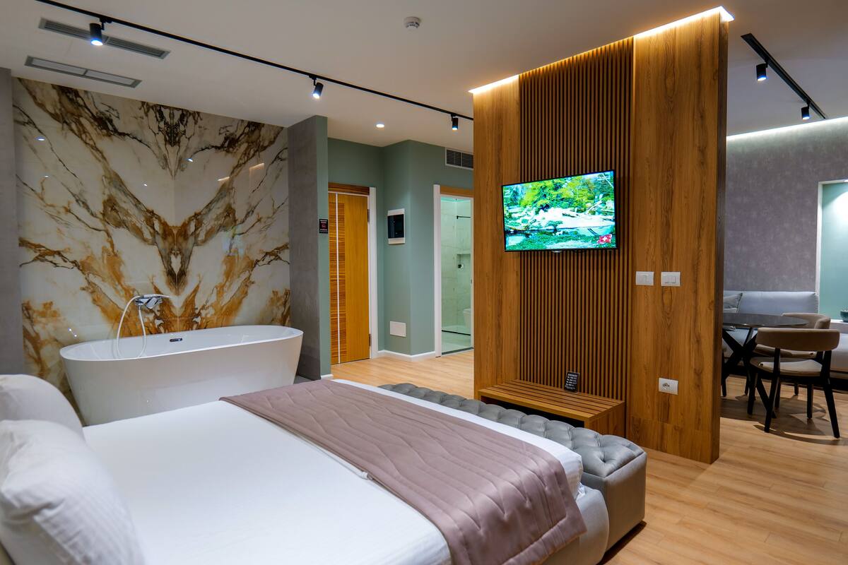 A modern hotel room with a large bed, a bathtub, a television, and a wooden panel wall.