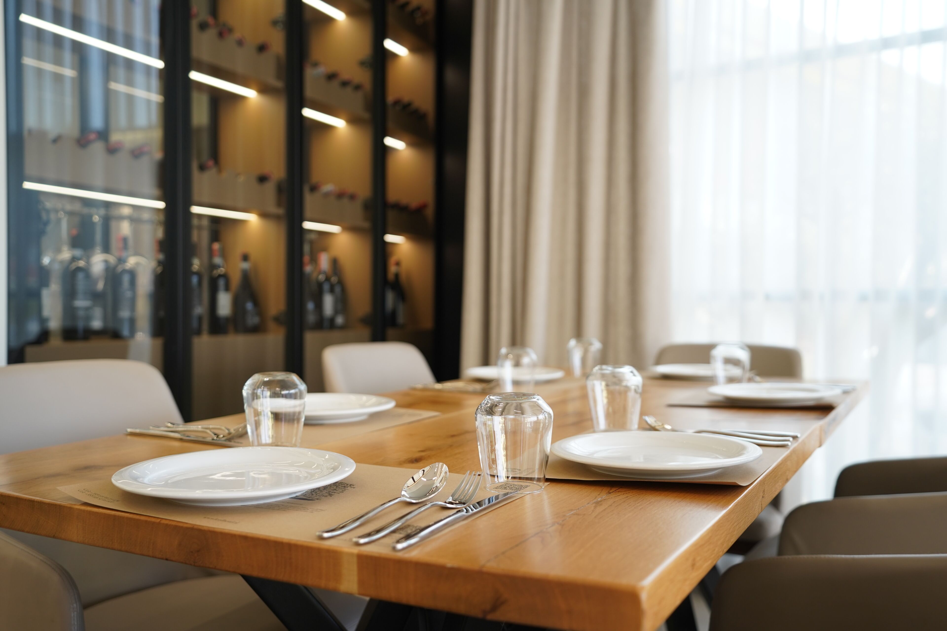 A dining table set with plates, cutlery, and glasses, with a modern interior and a view of bottles in the background.