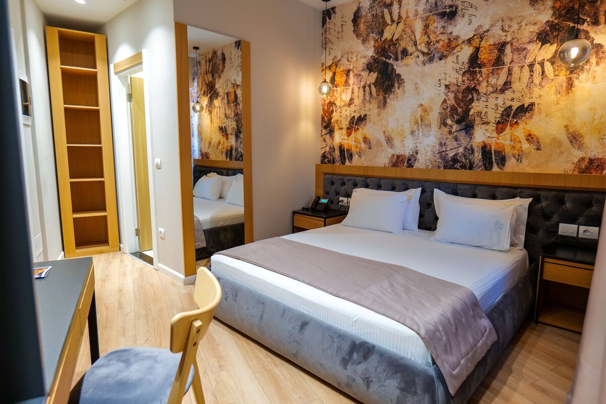 Een hotelkamer met een groot bed, nachtkastjes, een bureau en een stoel. Er is een garderobe en een spiegel aan de muur.