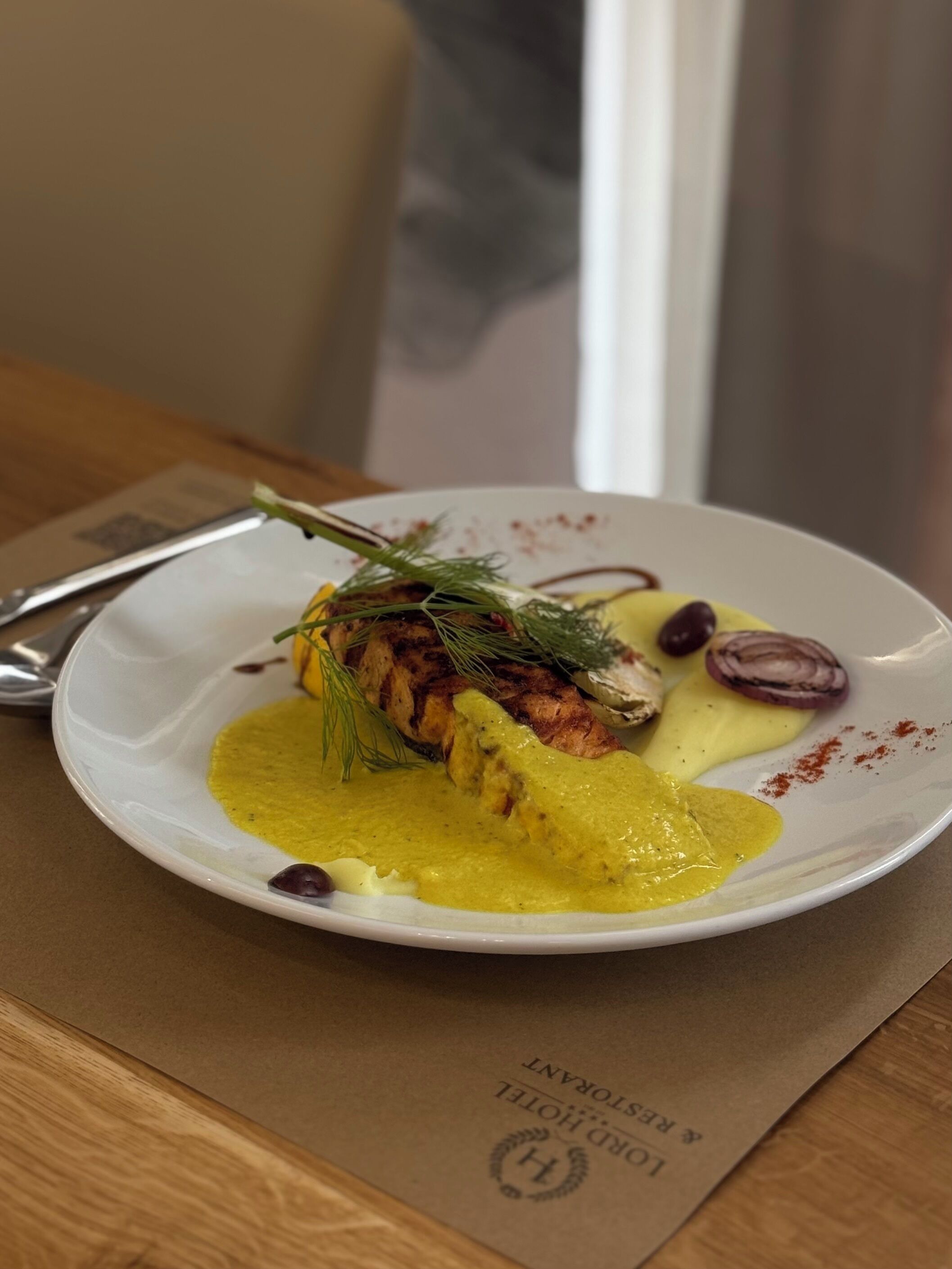 Un plato de comida con un filete de pescado, adornado con eneldo, acompañado de salsa amarilla y rodajas de cebolla roja.