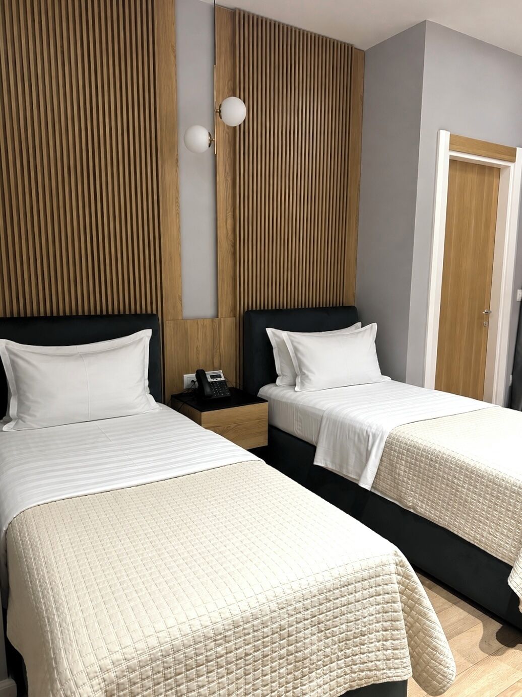 Quarto de hotel com duas camas, parede de painel de madeira, mesa de cabeceira com telefone e uma porta.