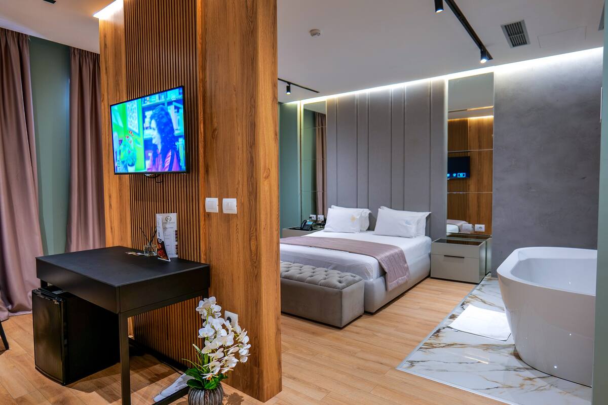 Een moderne hotelkamer met een houten accentmuur, een flatscreen televisie, een bed in grijs- en wit kleurenschema, een groot ligbad en een klein bureau met een bloevas.