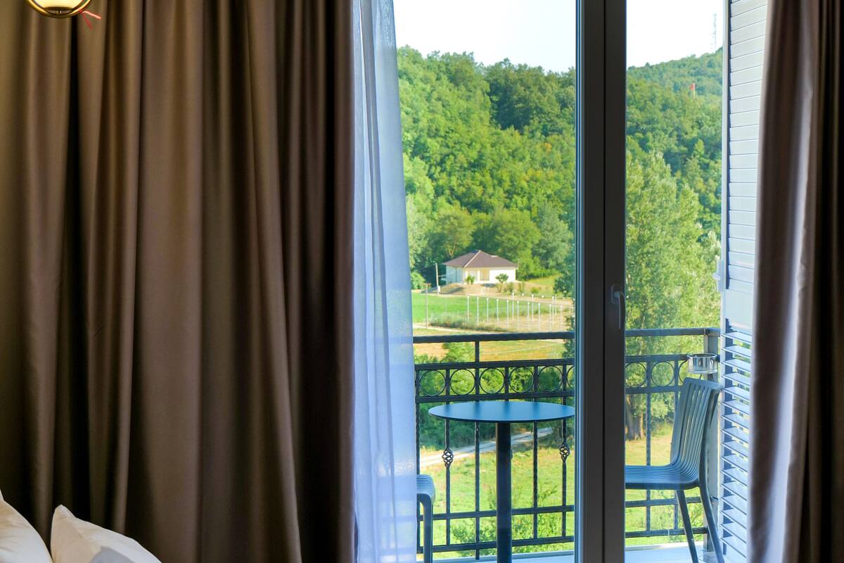 Een hotelkamer met een bed, een balkon met een tafel en stoelen, en uitzicht op een groen landschap.