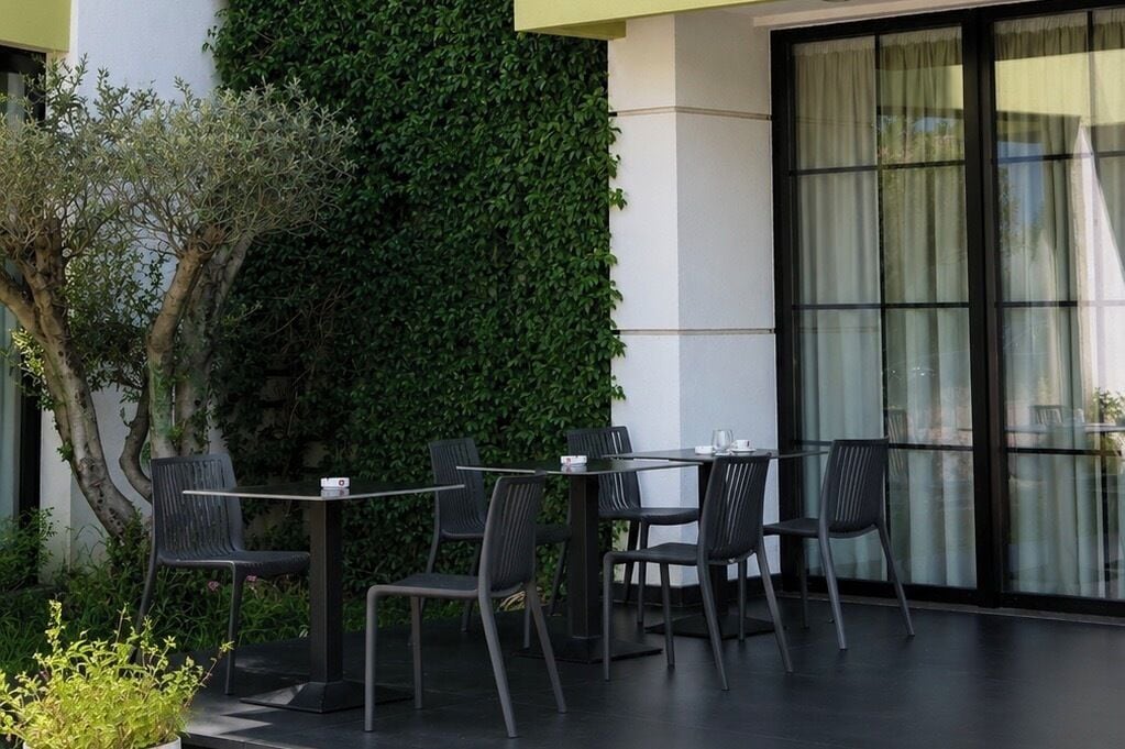 An outdoor seating area with tables and chairs, a large window with curtains, and a wall covered in greenery.