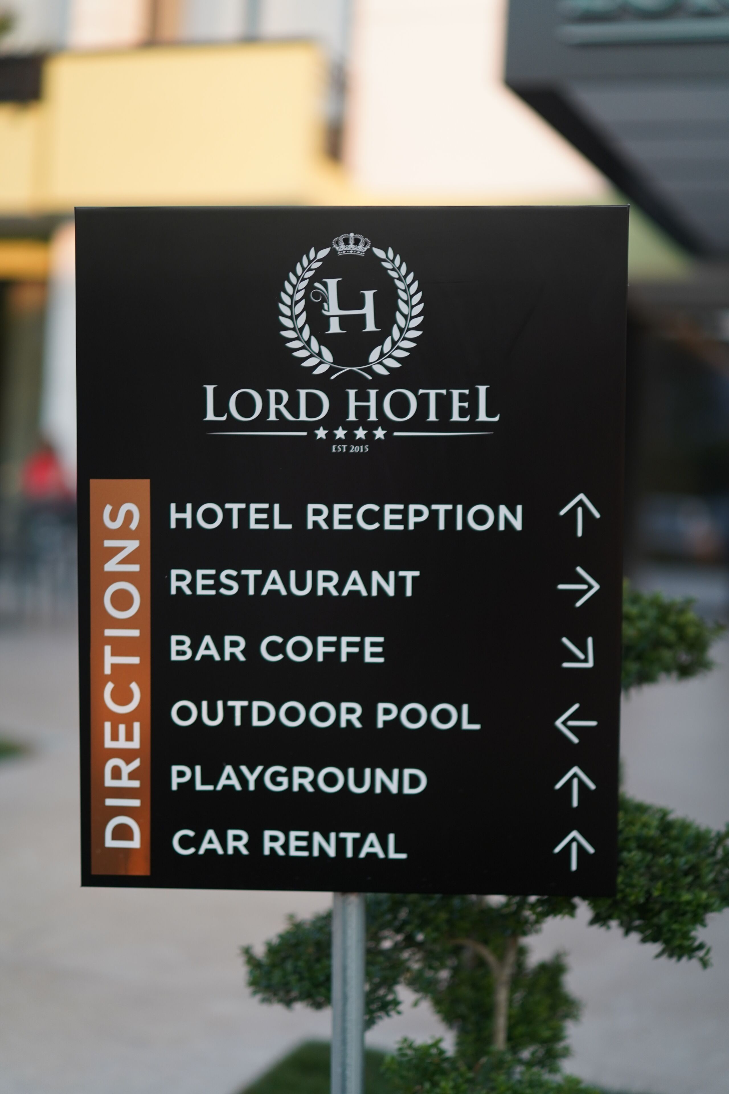 Panneau de signalisation indiquant la direction de l’hôtel Lord et mentionnant les services proposés : réception, restaurant, bar-cafétéria, piscine extérieure, aire de jeux et service de location de voitures.