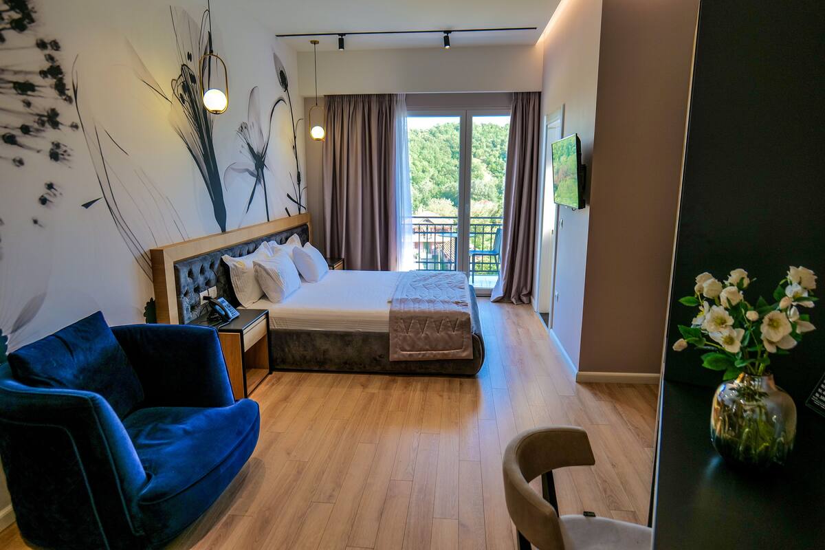A modern hotel room with a large bed, a blue sofa, a small table, and a balcony with a view of greenery.