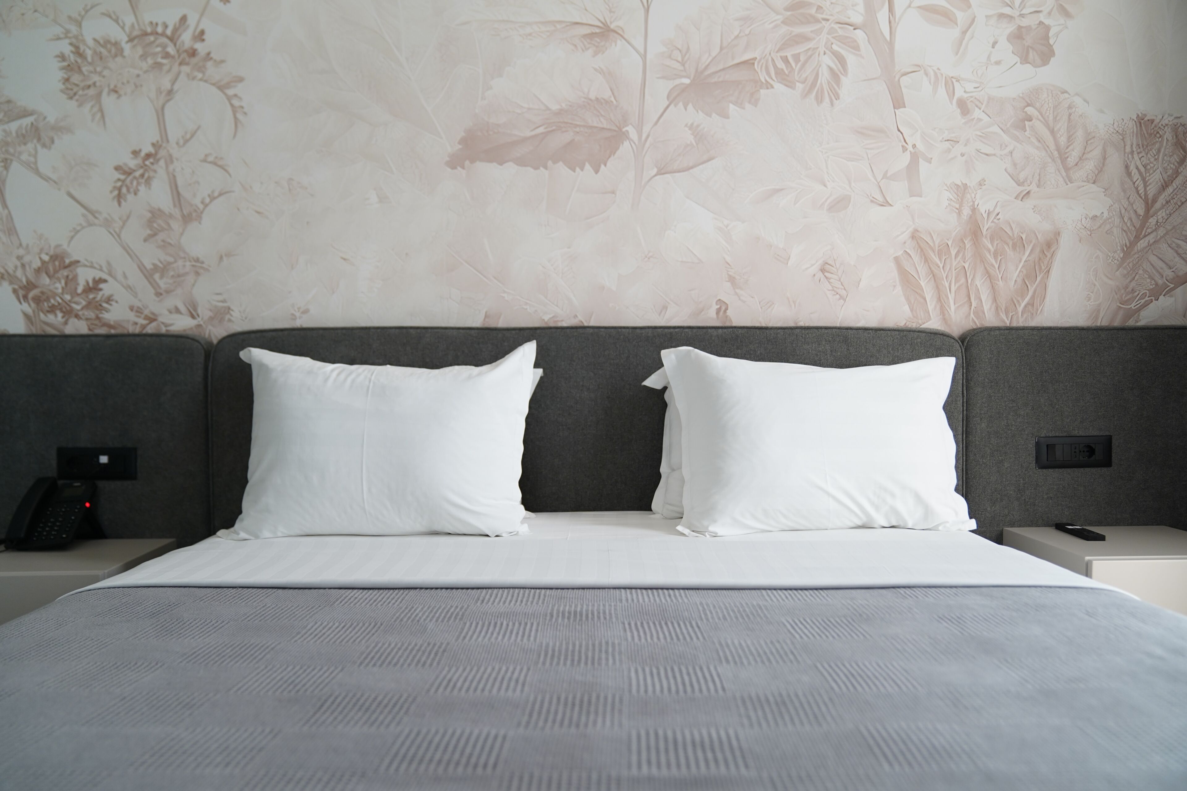 A hotel room with a large bed, two white pillows, a grey headboard, and a floral wallpaper.