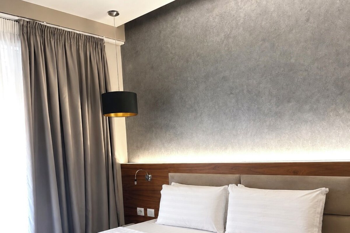 A hotel room with a large bed, a headboard, a wall-mounted lamp, and a window with curtains.