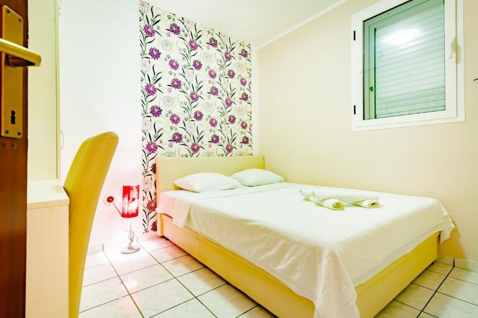 Une chambre à coucher avec un lit, une chaise, une fenêtre et un mur recouvert d’un papier peint à motifs floraux.