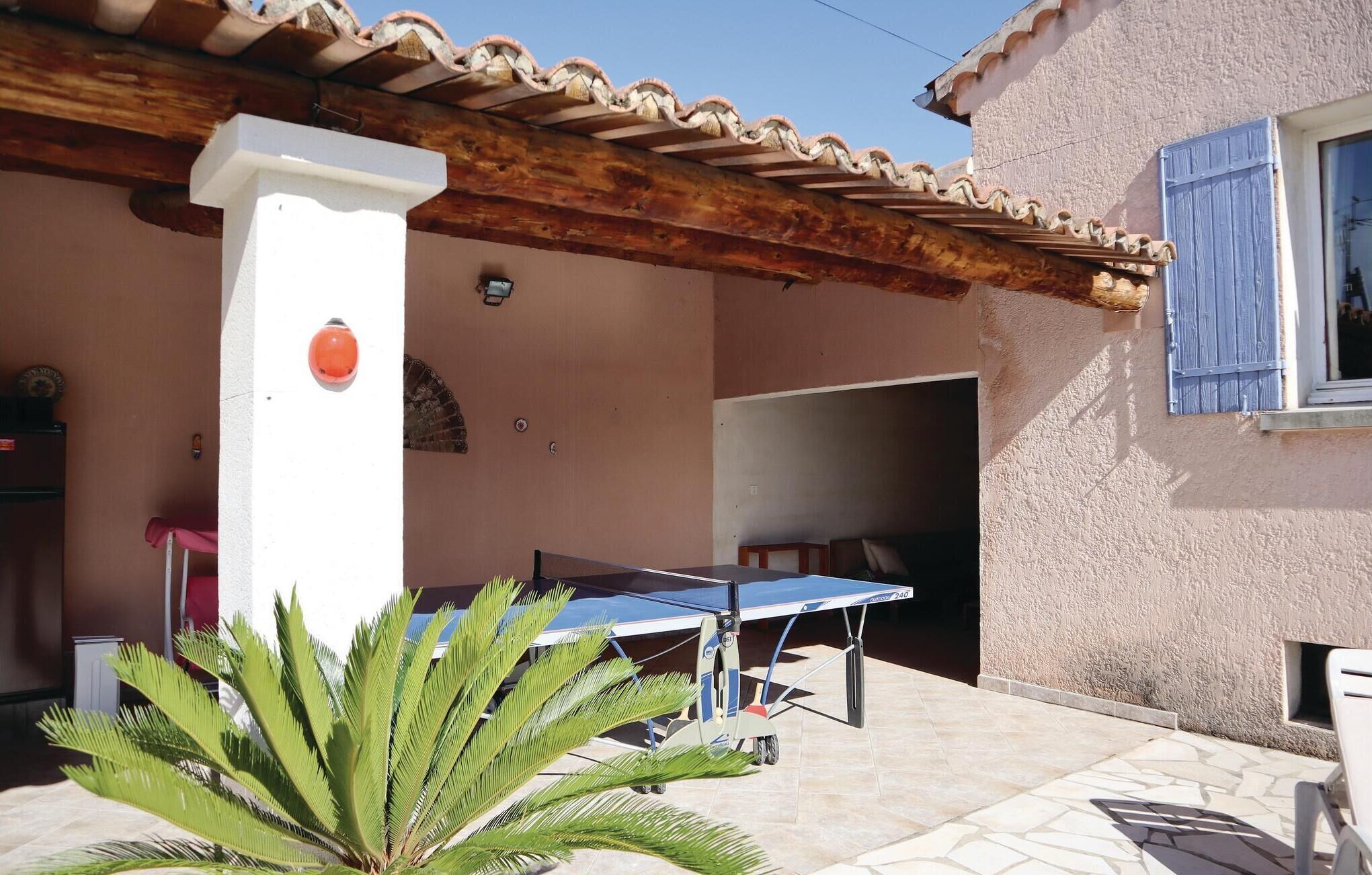 A covered outdoor area with a table and chairs, a palm tree, and a building with a tiled roof and a window with blue shutters.