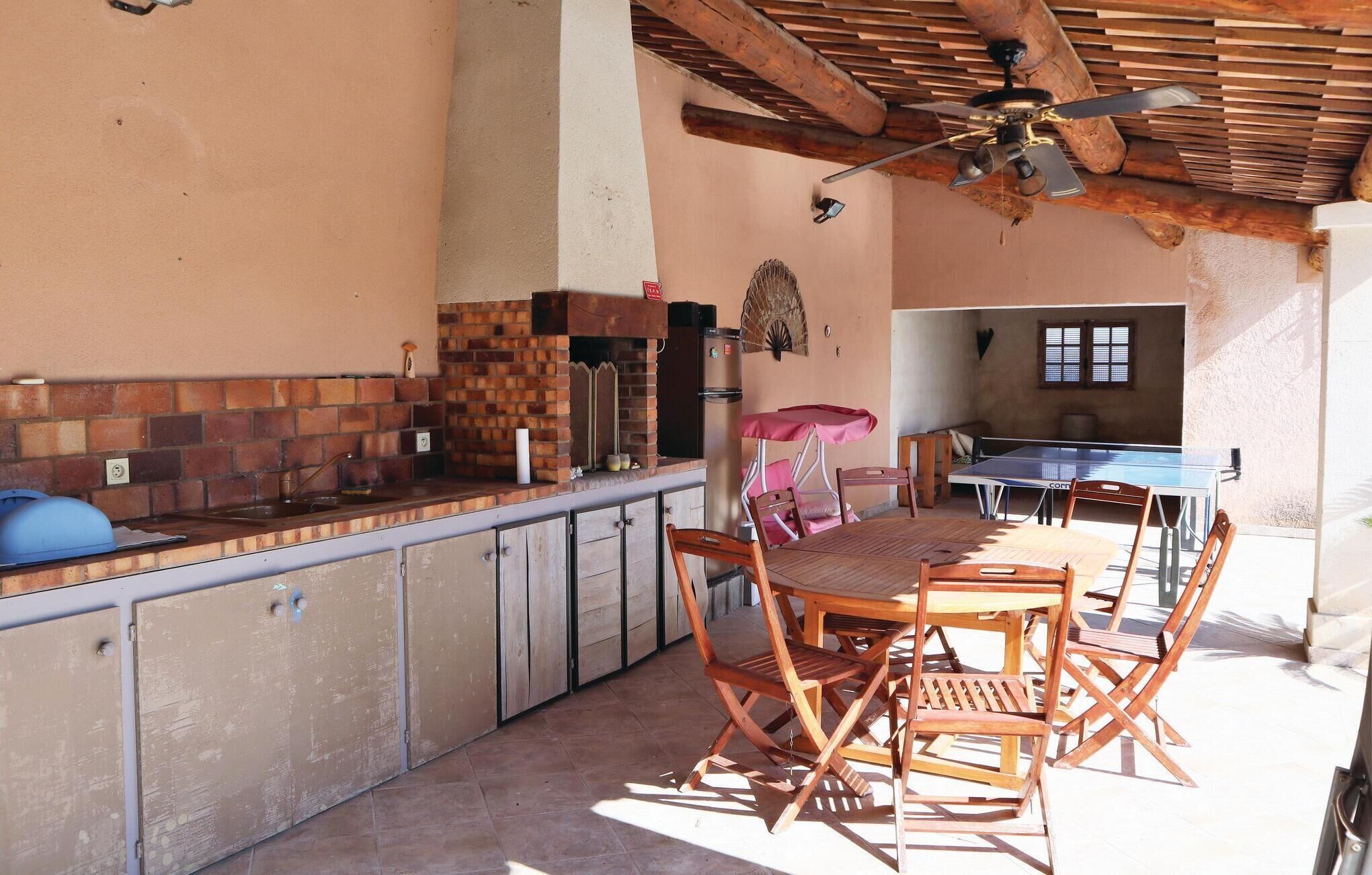 An outdoor kitchen area with a brick backsplash, a sink, and a ceiling fan. There is a dining table with chairs and a ping pong table in the background.