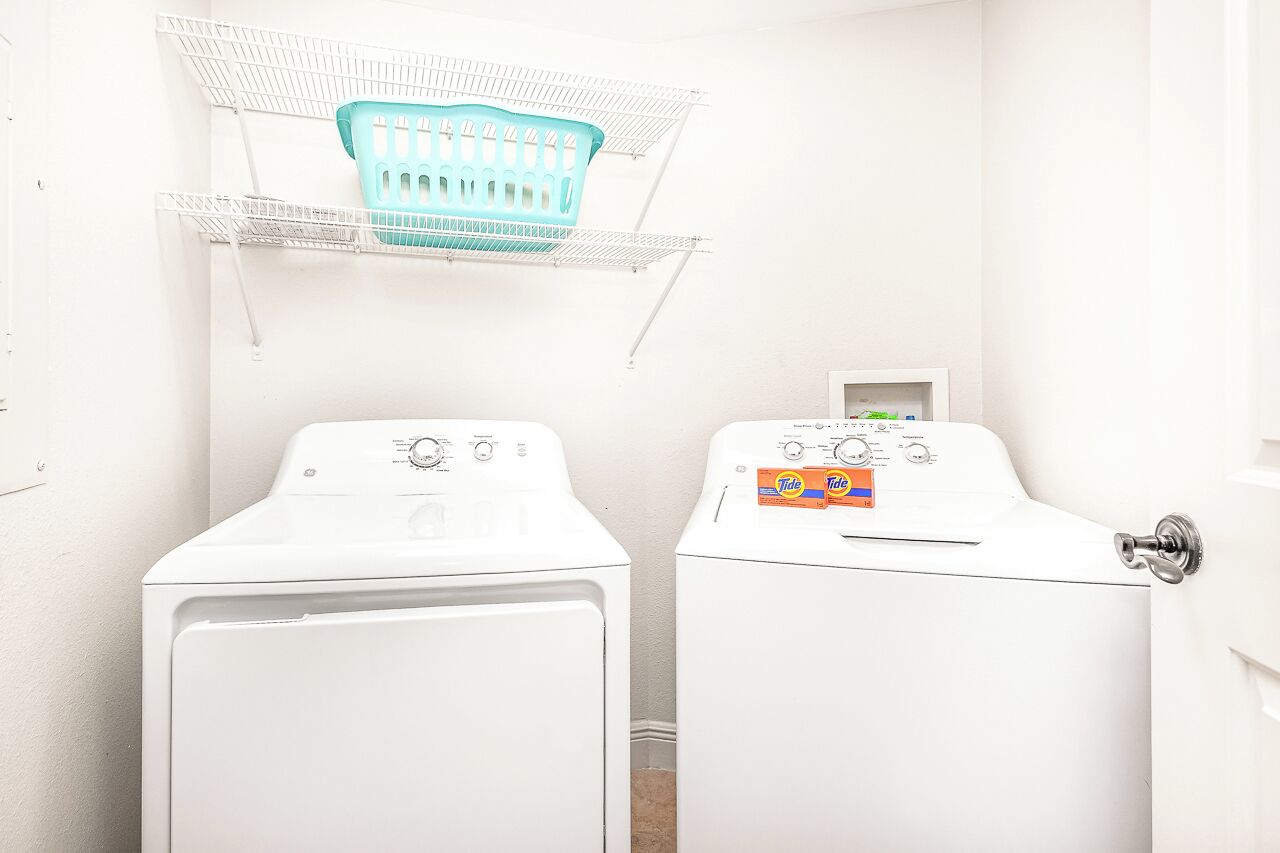 A laundry room with two white front-loading washing machines, a teal laundry basket on a white shelf, and a Tide detergent box on one machine.