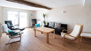 Un salón espacioso con suelo de madera, un sofá negro con cojines estampados, un sofá blanco con un cojín azul, una mesa de centro de madera y un estufa de leña negra.