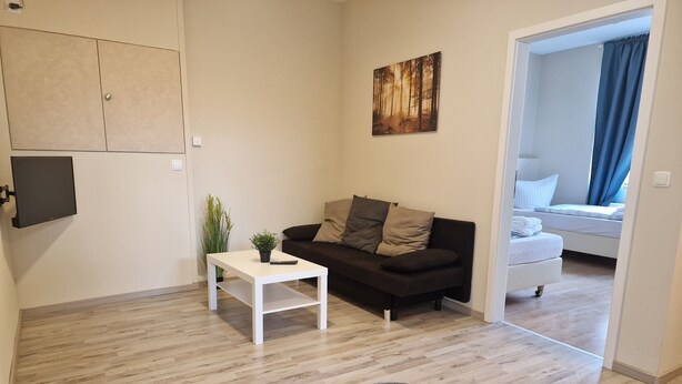 Un salon moderne avec un canapé, une petite table blanche ornée d’une plante, un tableau accroché au mur et une vue sur une autre pièce où se trouve un lit.