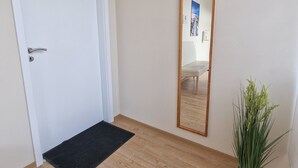 Une pièce avec un plancher en bois, une porte blanche, un paillasson noir, un miroir et une plante en pot.