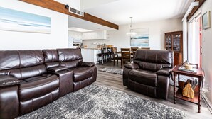 A living room with a brown leather sofa and armchair, a dining area with a wooden table and chairs, and a kitchen in the background.