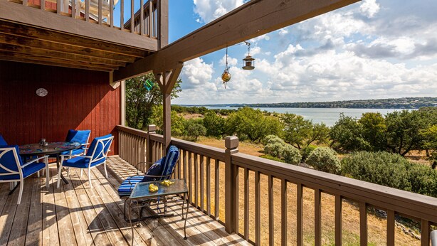 A wooden deck with blue chairs and a table, overlooking a lake and trees.