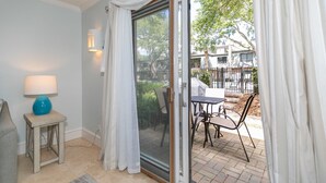 Et værelse med en skydedør, der fører ud til en udendørs terrasse. Der er et lille bord med stole og en lampe på et natbord.