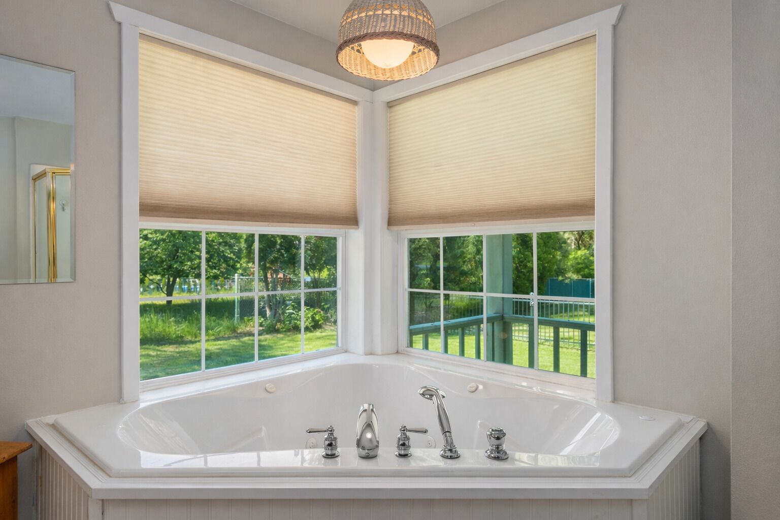 Ein Badezimmer im Eckbereich mit einer großen Badewanne, zwei Fenstern mit Jalousien und Blick auf eine grüne Außenanlage.